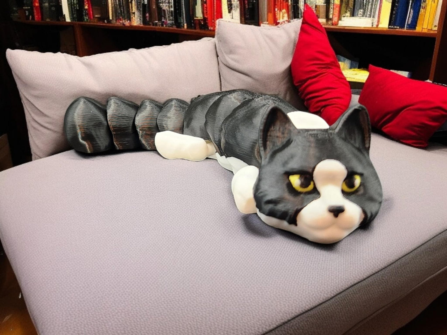 3D printed articulated cat figurine in gray and white lying on a purple couch with books and red pillows in the background