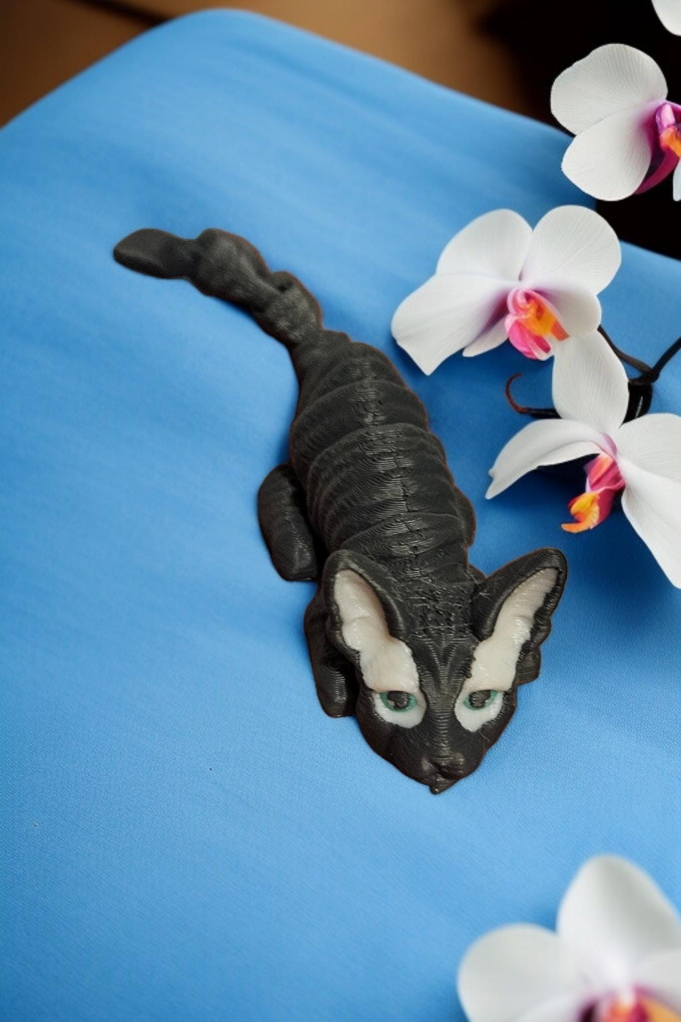 Grey 3D printed articulated cat figurine displayed on blue surface near white orchids