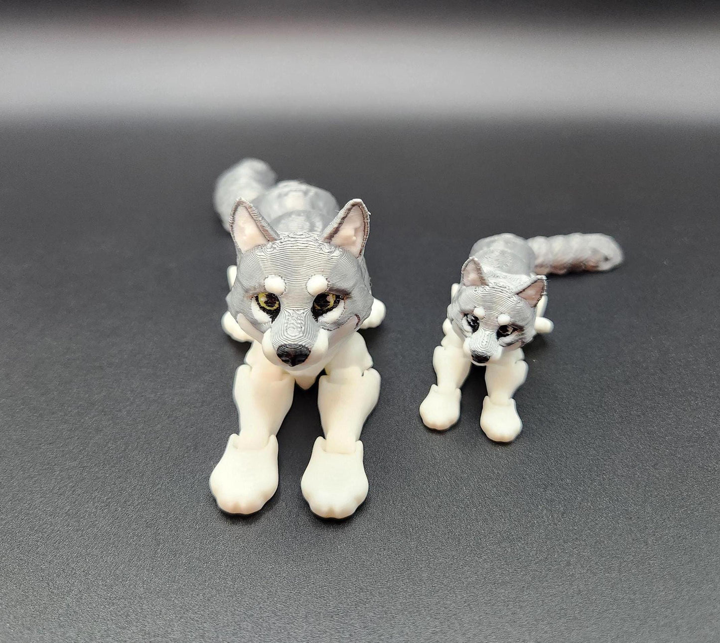Two 3D printed realistic wolf figurines in gray and white lying down on a black surface