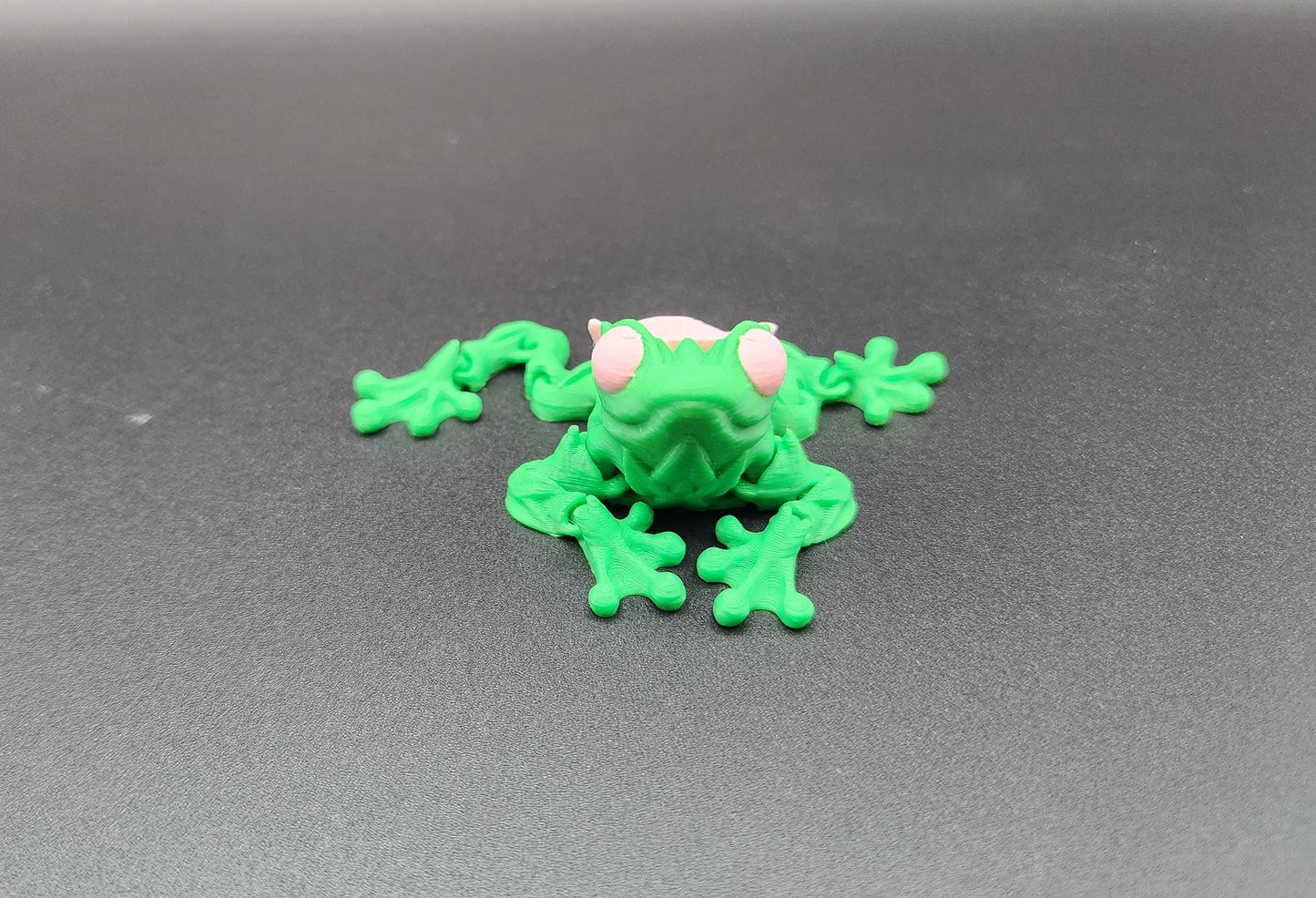 Green 3D printed articulated rose frog toy with pink eyes on a gray surface