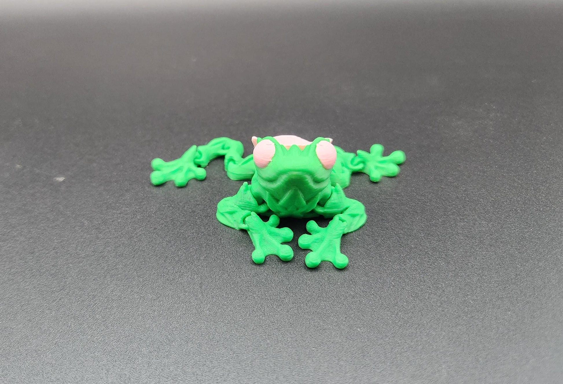 Green 3D printed articulated rose frog toy with pink eyes on a gray surface