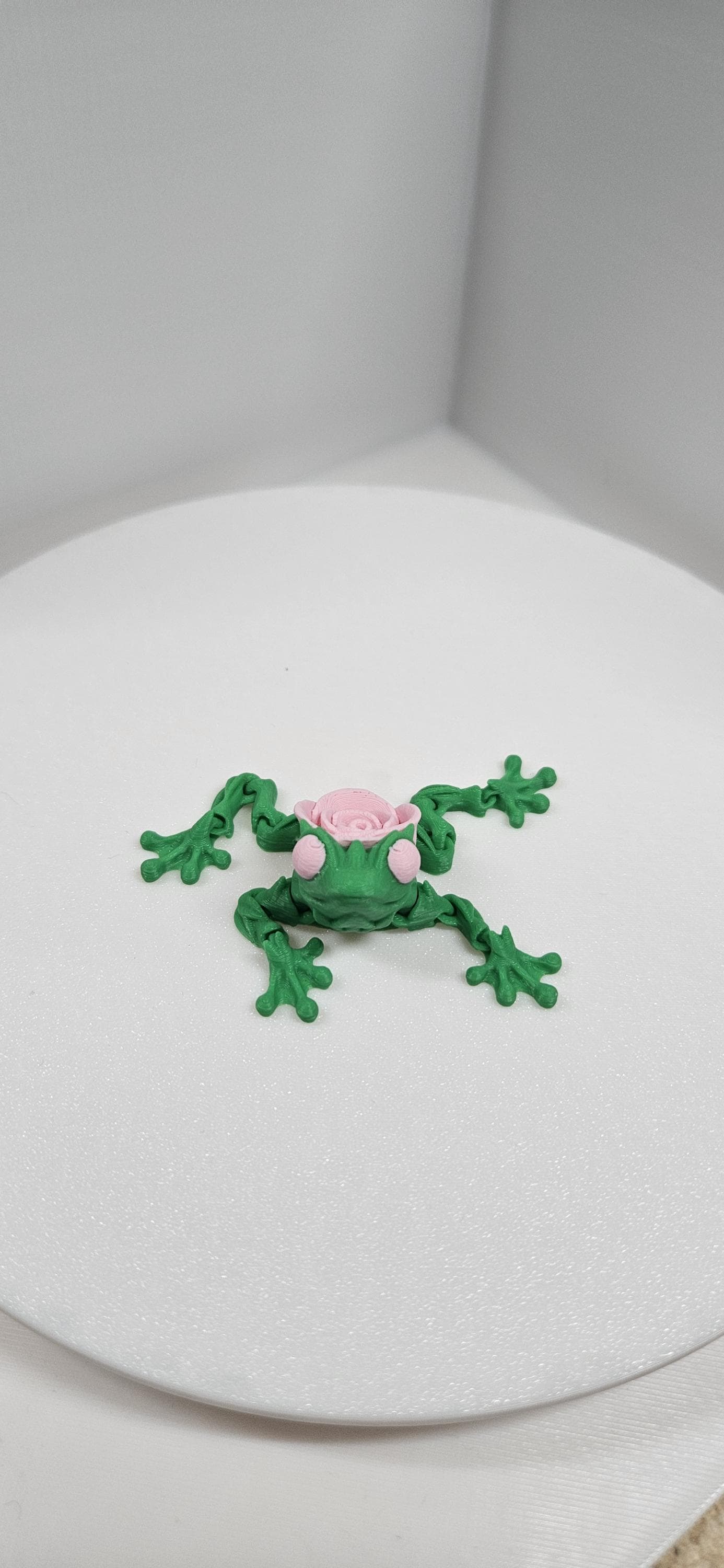 Green 3D printed articulated rose frog with pink rose detail on its back displayed on a white surface