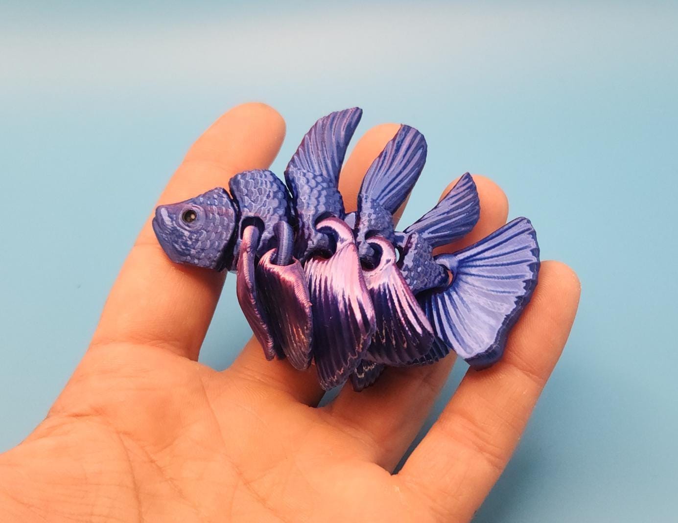 Blue and purple 3D printed betta fish toy held in a hand on a light blue background