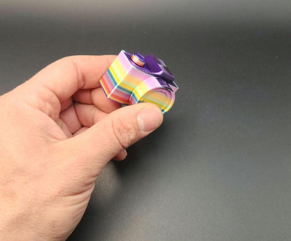Hand holding a colorful 3D printed camera fidget keychain with a rainbow pattern and purple details