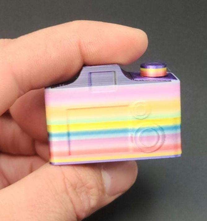 Hand holding a colorful 3D printed camera fidget keychain with rainbow stripes on a dark background