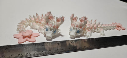Pair of 3D printed cherry blossom baby dragons with pink floral details and blue eyes on white background
