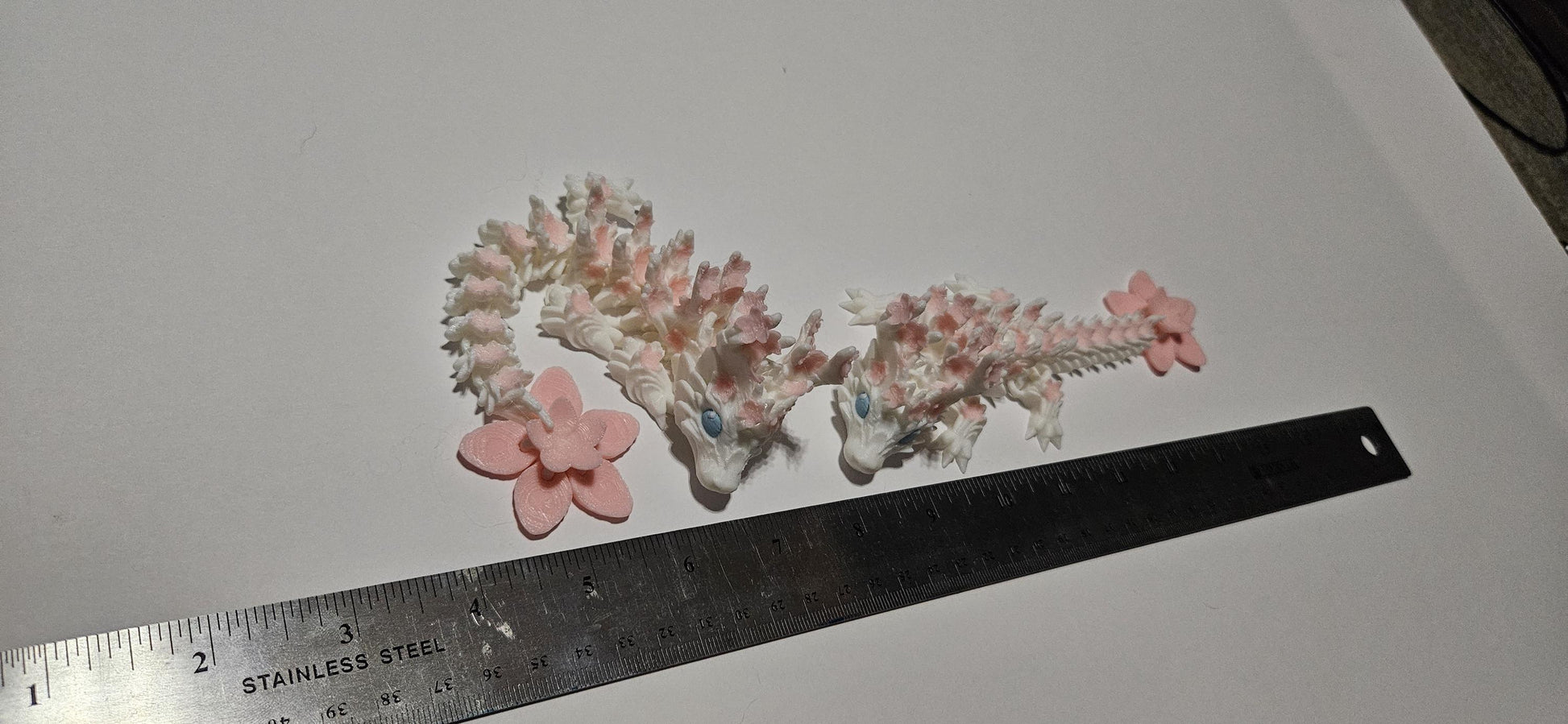 Two small 3D printed cherry blossom baby dragons with pink flowers next to a ruler for size reference