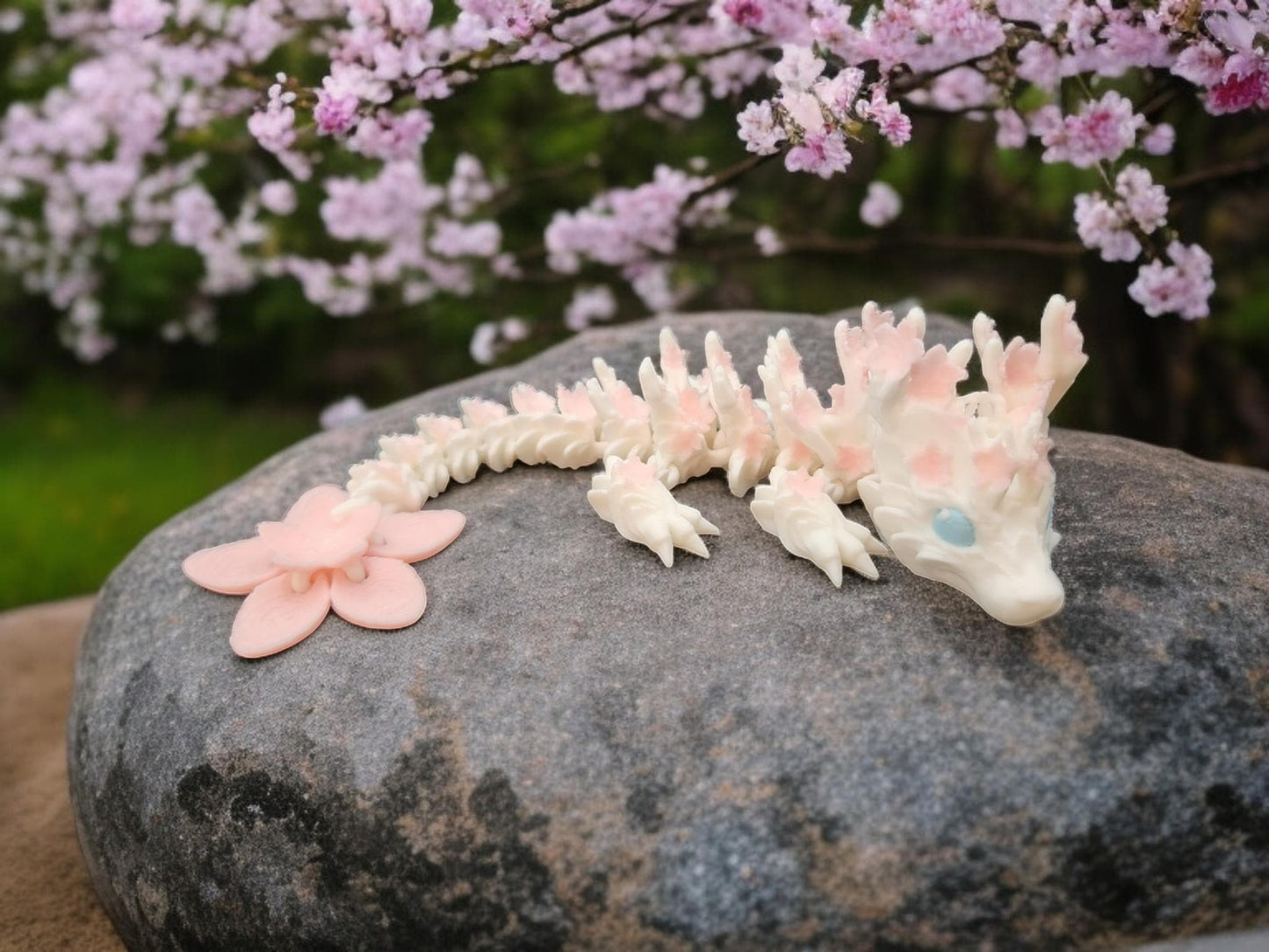 3D printed cherry blossom baby dragon figurine in soft pink and white colors resting on a rock with cherry blossoms in background