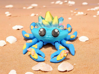 Blue 3D printed crystal crab with yellow and pink accents on sandy beach with seashells