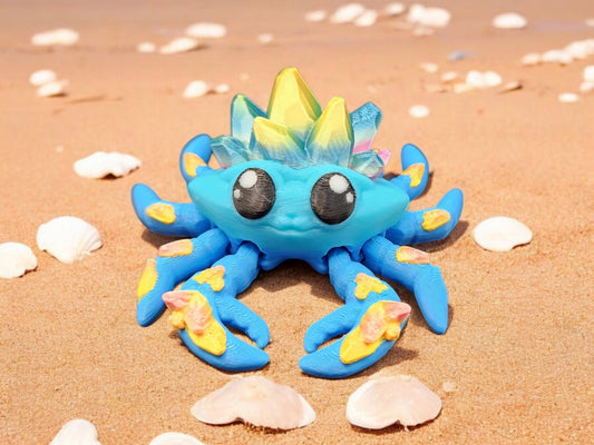 Blue 3D printed crystal crab with yellow and pink accents on sandy beach with seashells