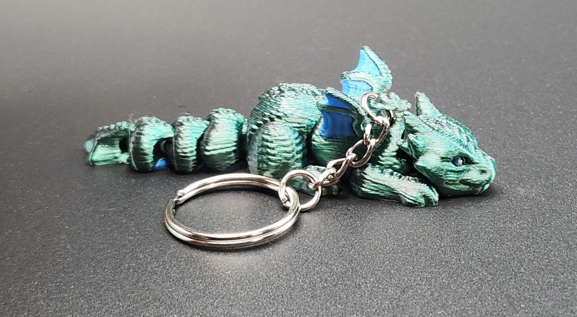 3D printed dragon keychain in green and blue colors with detailed scales and wings on black surface