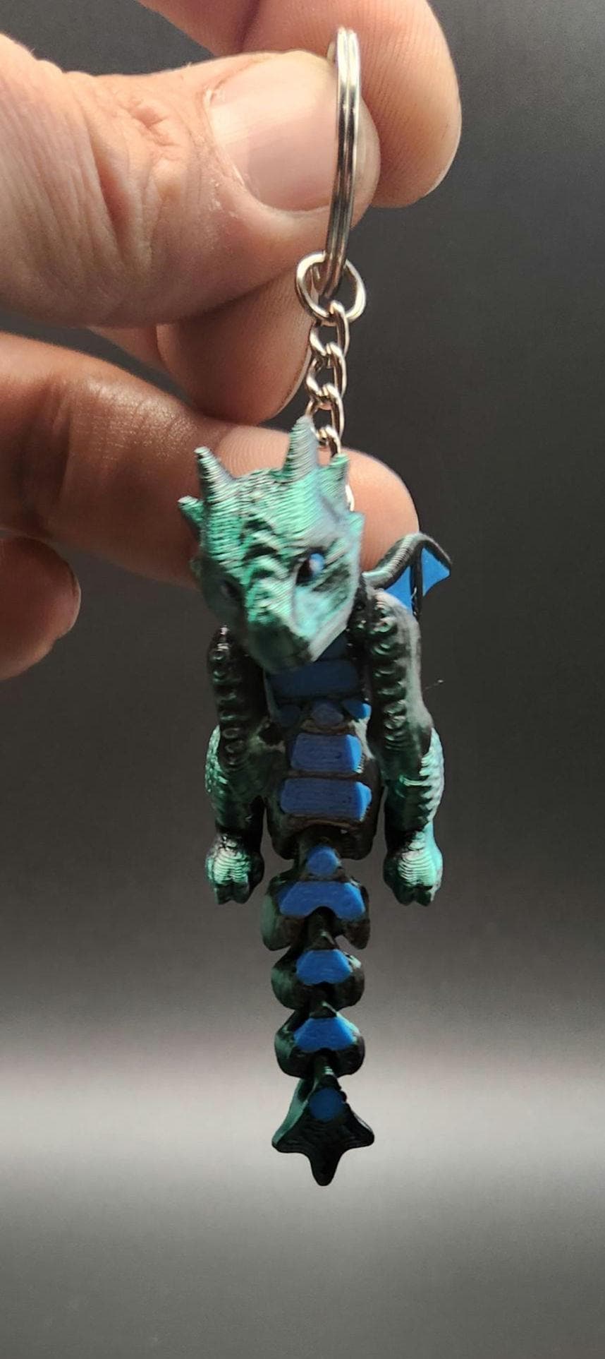 Hand holding a 3D printed dragon keychain with blue and green scales and metal keyring