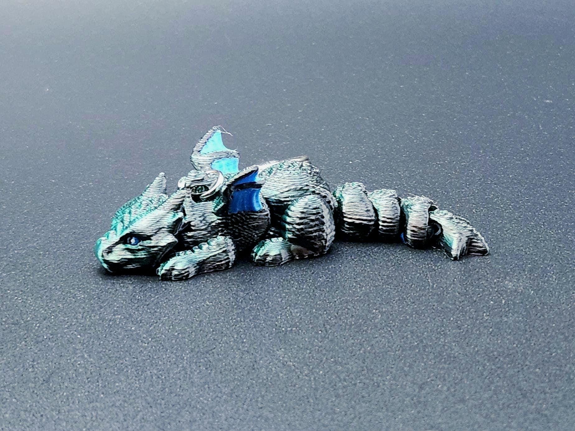Blue and gray 3D printed dragon keychain with detailed scales and wings lying on a dark surface