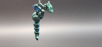 Green and blue 3D printed dragon keychain hanging against a gray background with detailed wings and tail