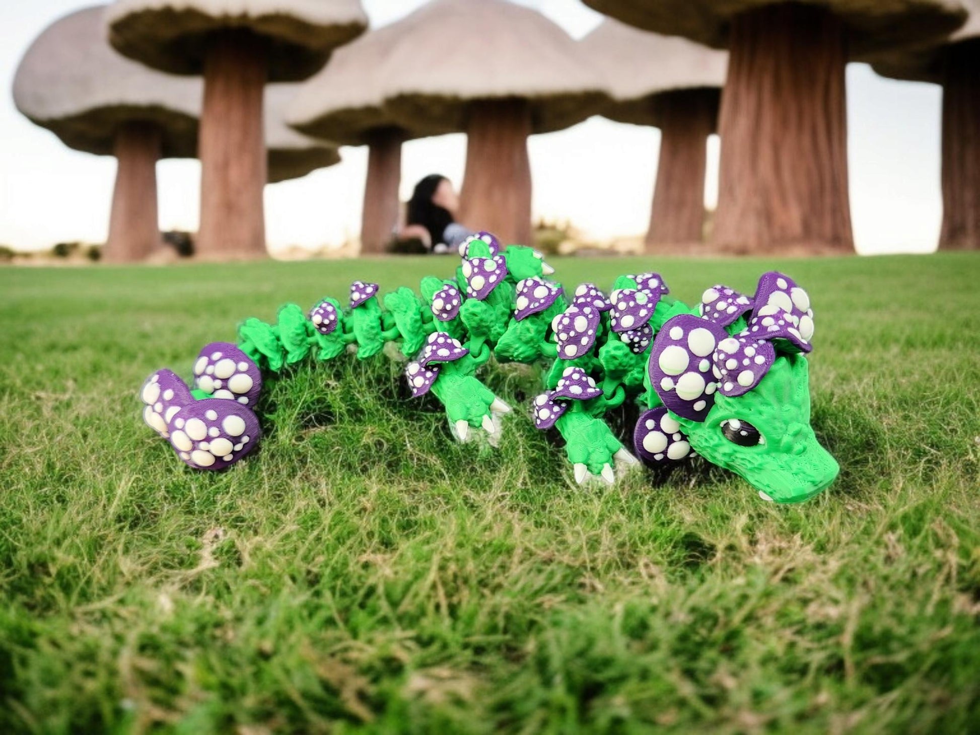 Green and purple 3D printed dragon mushroom toy with white spots displayed on grass with large mushroom sculptures in background