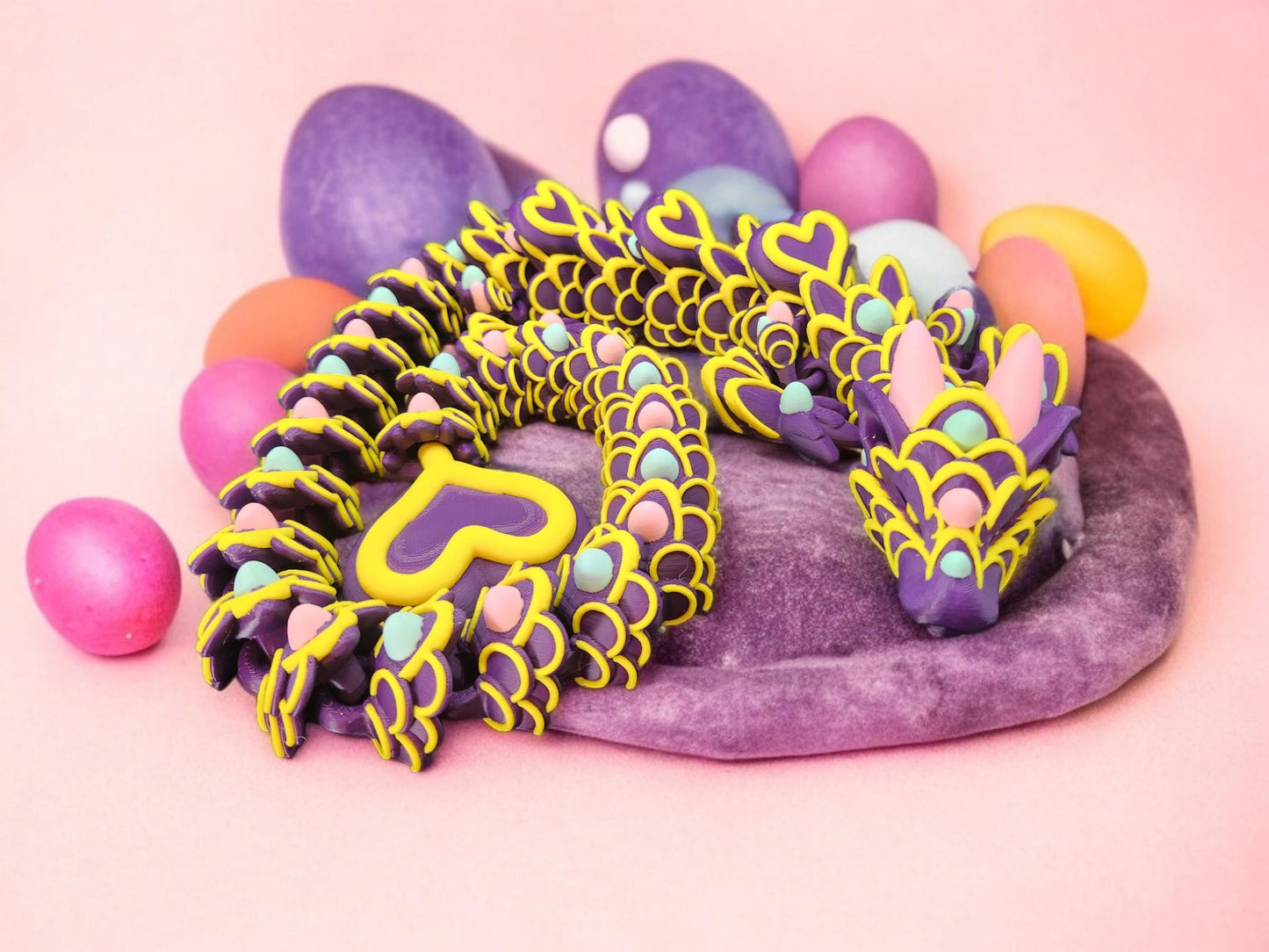 Colorful 3D printed Easter dragon in yellow and purple surrounded by pastel Easter eggs on a soft purple mat