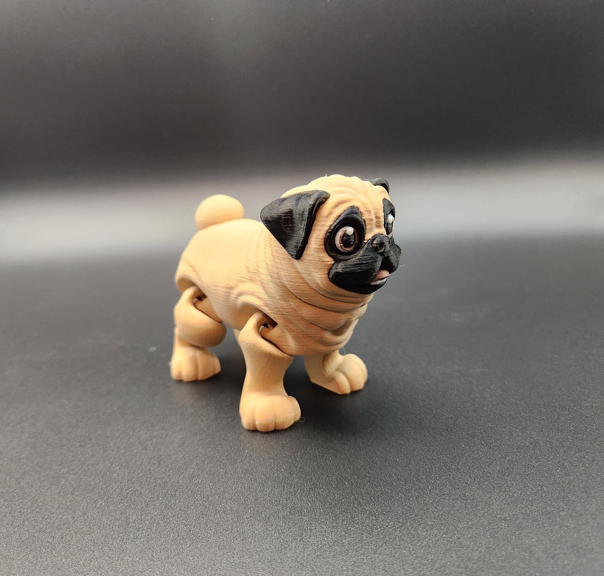 Cute 3D printed pug toy in beige and black with articulated legs on a dark background