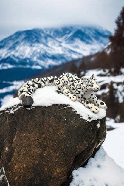 3D printed snow leopard figurine lying on a snow-covered rock with snowy mountains in the background