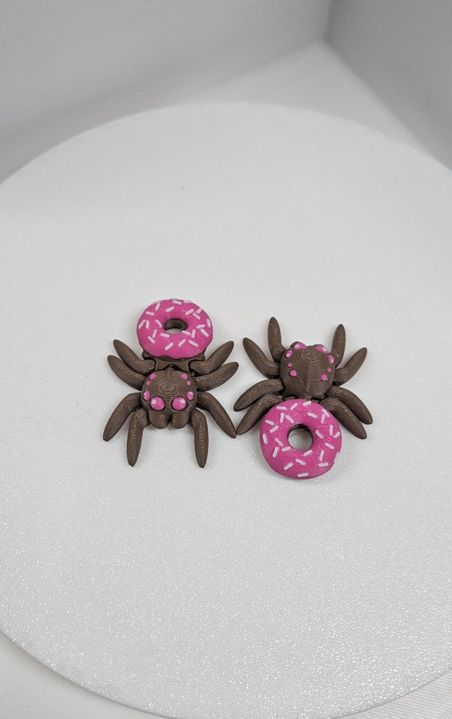 Two 3D printed spider fidget toy figures with pink donut designs on a white surface