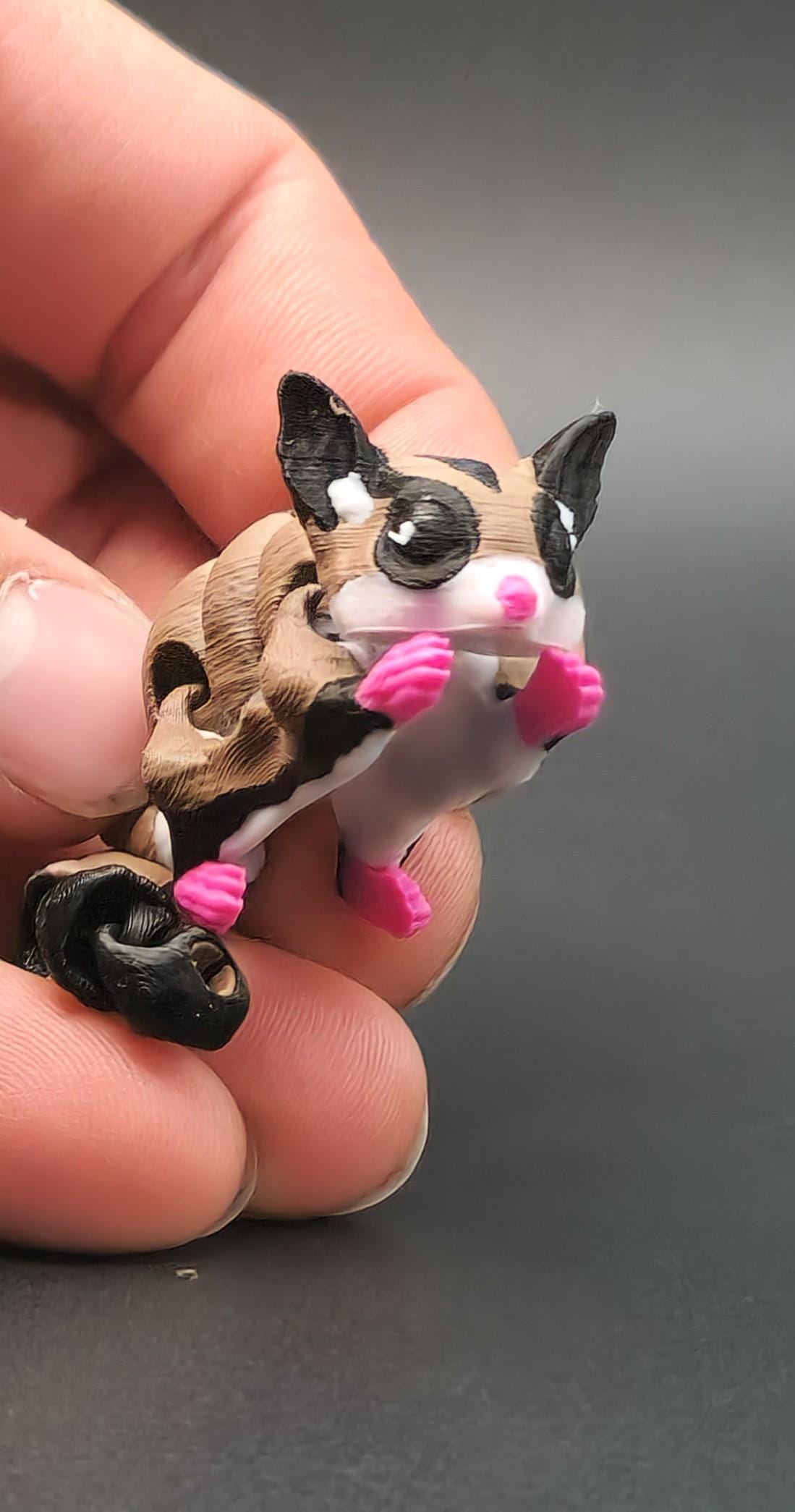 Hand holding a 3D printed sugar glider keychain with detailed pink paws and black striped design