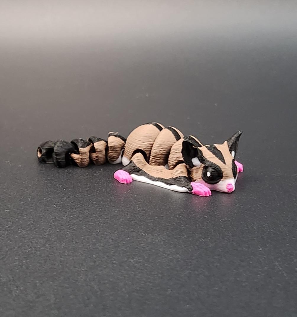 Close-up of a 3D printed sugar glider keychain in beige, black, and pink colors on a black surface