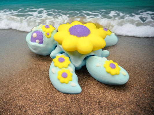 3D printed sunflower turtle figurine with yellow and purple flowers on sandy beach near ocean waves