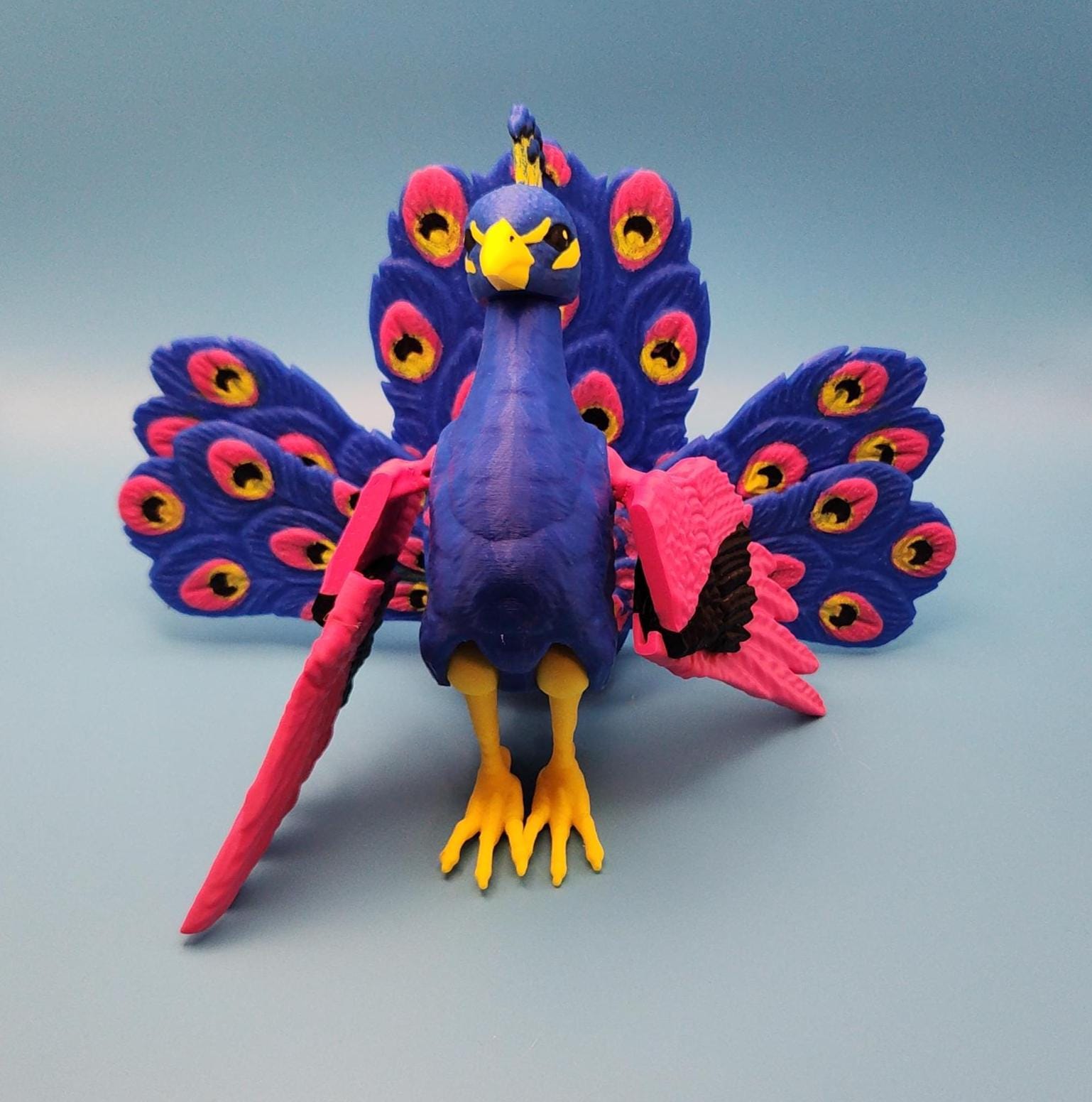Colorful articulated bird desk decor of a peacock with blue, pink, yellow, and black details on a gray background
