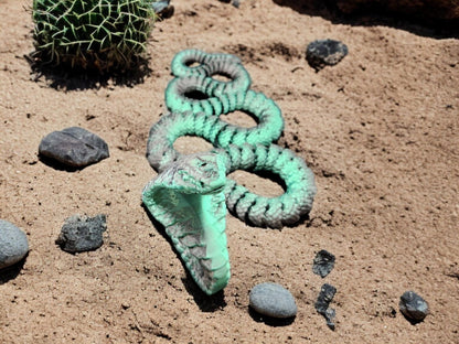 Green articulated king cobra toy posed on sandy surface with small rocks and cactus in background