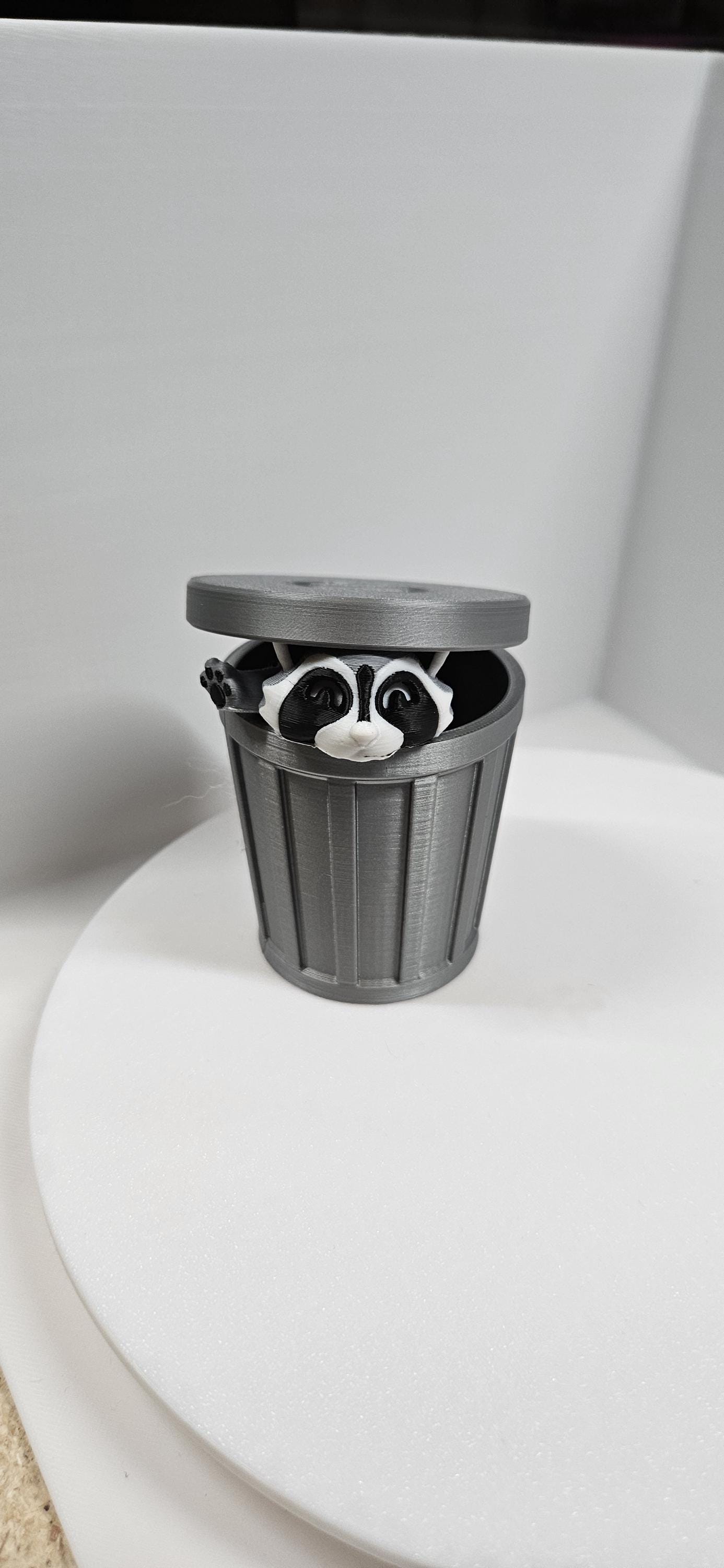 Articulated raccoon fidget figure peeking out of a gray trash can with a partially open lid