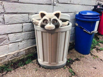 Articulated raccoon fidget figure peeking out of a gray trash can outdoors near a blue recycling bin