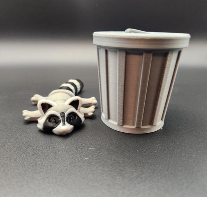 Small articulated raccoon fidget figure lying next to a gray trash can model on black surface